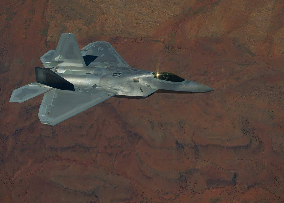 F-22 Raptor in flight with afterburners glowing, seen from below against a clear sky