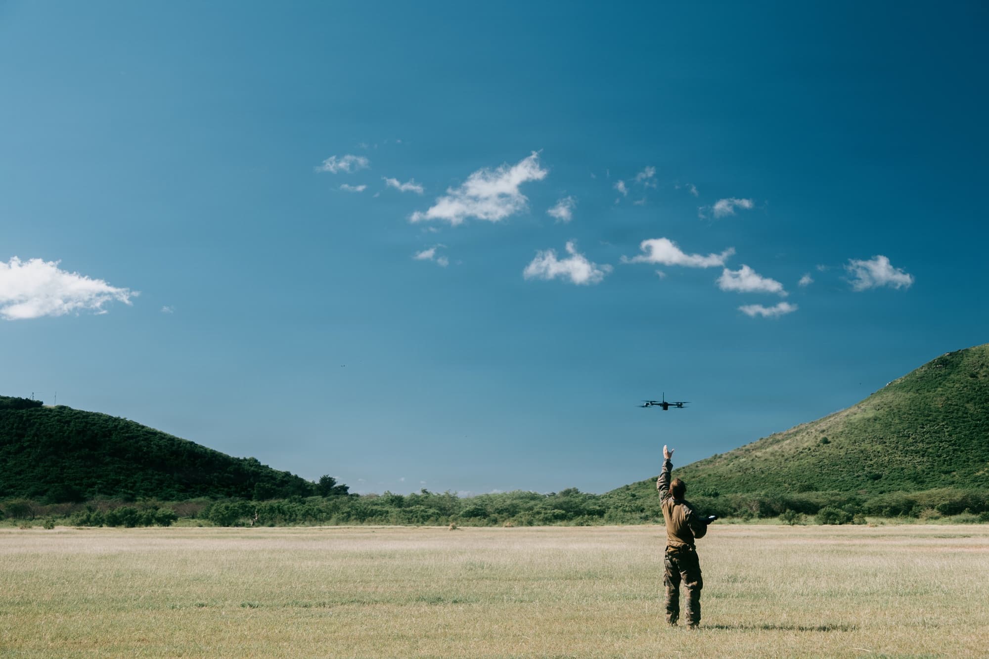 A military FPV drone trailing a thin fiber-optic cable during flight over terrain