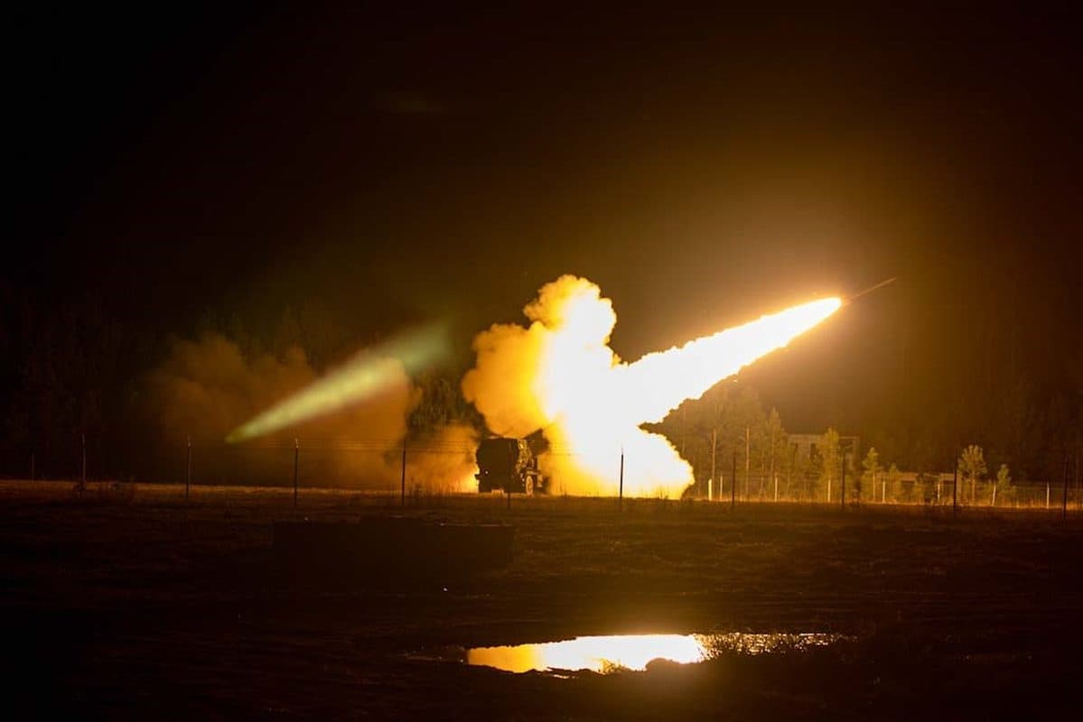 HIMARS launching a rocket during a night live-fire exercise, with dramatic exhaust flame and smoke trail illuminating the sky