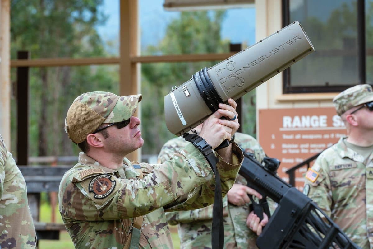 Military operator controlling an FPV drone in a forested environment, the drone hovering overhead during a NATO training exercise
