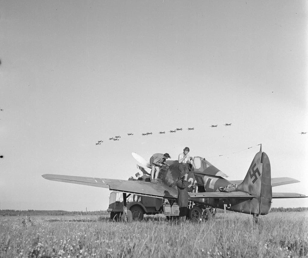 Fw 190A on the ground with crew working around the cockpit area, aircraft formation visible overhead