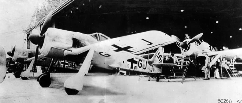 Multiple Focke-Wulf Fw 190 fighters inside a factory hangar with maintenance crews on ladders