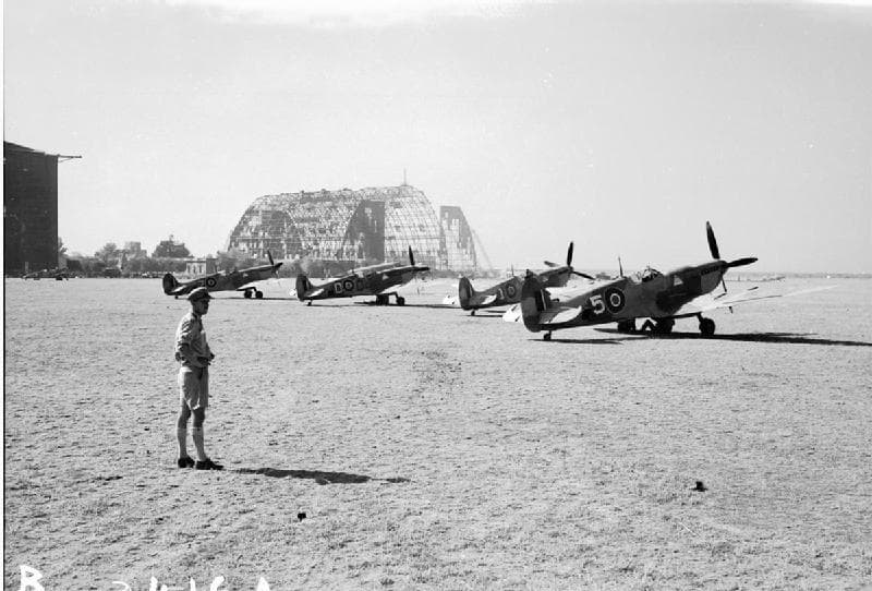 Multiple Spitfires parked at an airfield dispersal area with a damaged hangar in the background