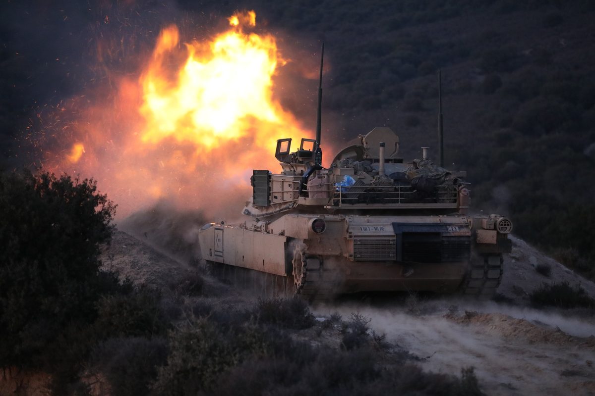 M1 Abrams tank firing at a target during live fire exercise in Greece with dust and muzzle blast visible