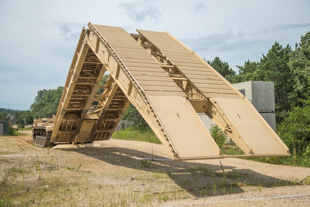 An Armored Vehicle Launched Bridge deploying its folding bridge span from the chassis of a modified tank hull, with the bridge extending forward over a gap