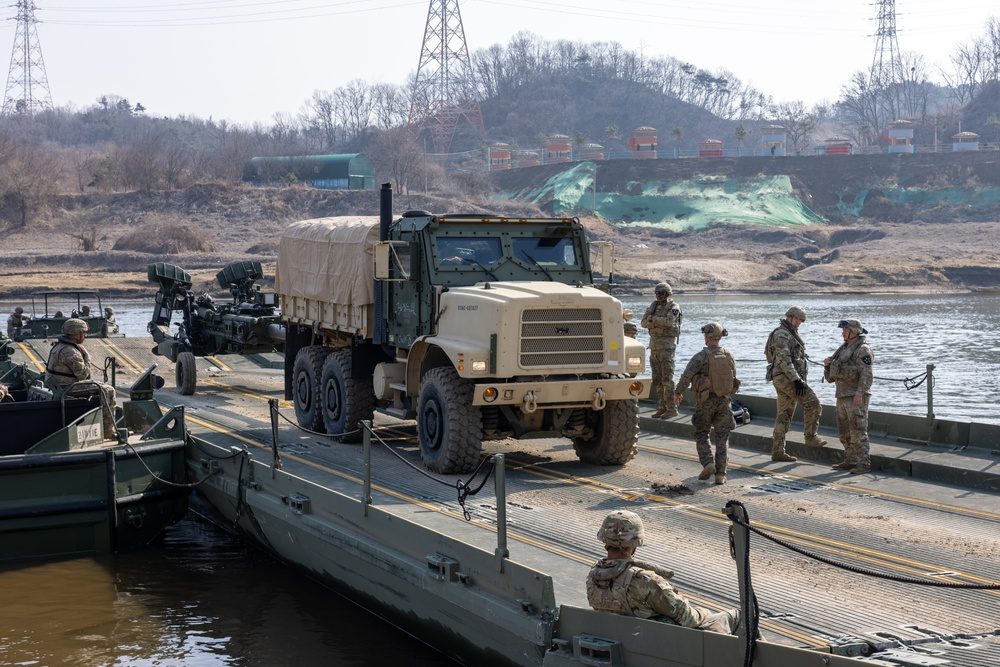 Improved Ribbon Bridge sections connected and floating on a river, forming a continuous roadway across the water with vehicles visible on the far bank