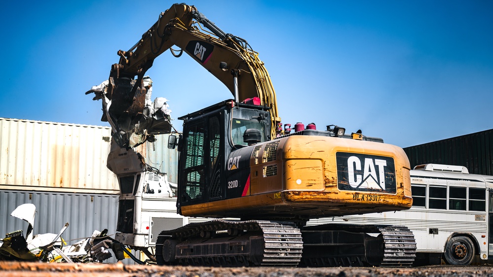 An excavator assigned to the Defense Logistics Agency tears apart a government owned vehicle