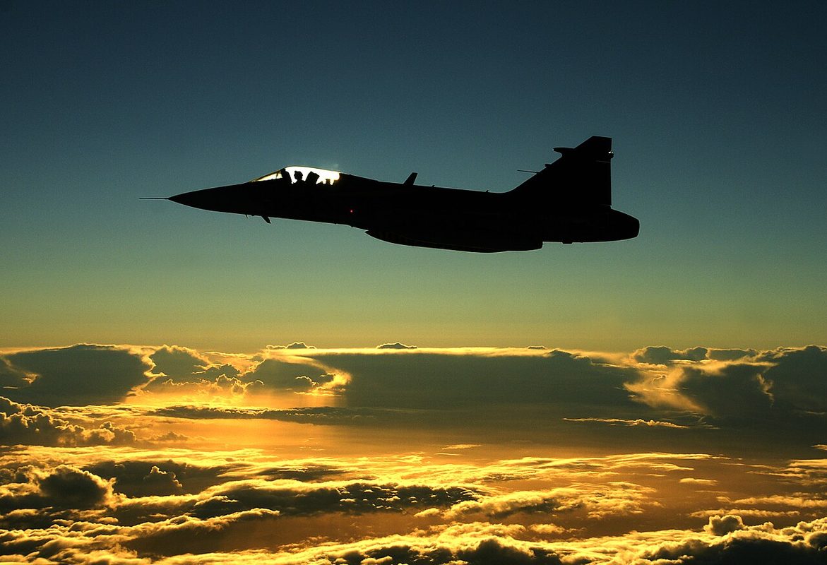 Saab Gripen fighter silhouetted in flight above clouds at sunset
