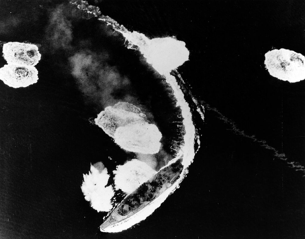 Yamato under aerial attack with bomb splashes and anti-aircraft fire visible around the ship